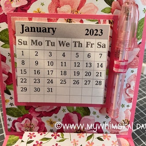 May include: A pink and white floral patterned card with a silver calendar for January 2023. The calendar has a grid with the days of the week labelled and the numbers 1 through 31. A pink pen is attached to the card.