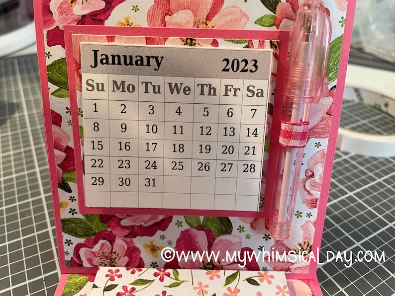May include: A pink and white floral patterned card with a silver calendar for January 2023. The calendar has a grid with the days of the week labelled and the numbers 1 through 31. A pink pen is attached to the card.