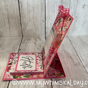 May include: A pink floral desktop calendar with a notepad that says "Hello" on the front. A pen is inserted into the calendar.