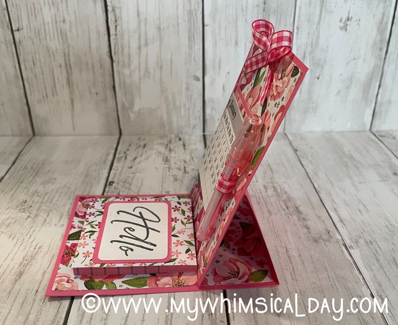 May include: A pink floral desktop calendar with a notepad that says "Hello" on the front. A pen is inserted into the calendar.