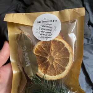 May include: A small, clear bag containing dried herbs and spices. The bag includes a dried orange slice, rosemary, cinnamon sticks, and a bay leaf with the word "HOPE" on it. A circular label reads "Yule Simmer Pot Bag".