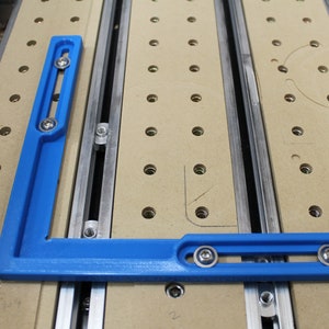 May include: A blue L-shaped woodworking jig with two sets of adjustable knobs. The jig is positioned on a pegboard surface with multiple holes.