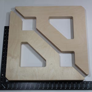 May include: A large, light-colored wooden square with cut-out geometric shapes. The square has a unique design with angled and rectangular openings. A ruler is placed along the bottom edge, providing scale. The wood has a natural grain.