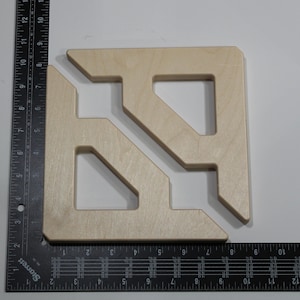 May include: A light-colored wooden square with a geometric design, featuring cut-out shapes. The square is placed on a black ruler with inch measurements. The wood has a natural, unfinished appearance.