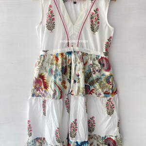 May include: White cotton dress with a V-neckline and a tie at the waist. The dress has a floral print in shades of pink, blue, yellow, and green. The dress is sleeveless and has a tiered skirt.