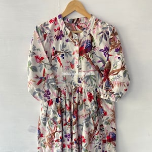 Puede incluir: Un vestido de algodón blanco con estampado floral con pájaros y flores de colores. El vestido tiene un frente abotonado y una falda fruncida. El texto "indiancottondress" está impreso en la parte delantera del vestido.