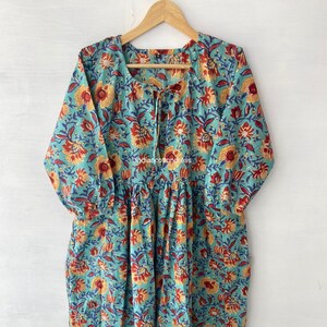 以下が含まれることがあります： 赤、黄色、青の色合いの花柄がプリントされたターコイズブルーの綿のドレス。ドレスはVネックで、ネックラインにリボンが付いています。ドレスは長袖で、ギャザーの入ったスカートです。