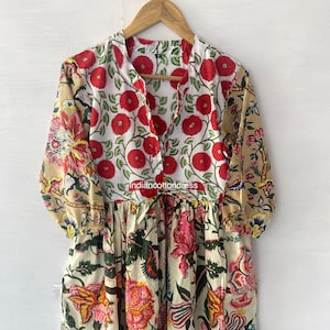 Könnte beinhalten: Ein weißes Baumwollkleid mit Patchwork-Design, das florale Muster in Rot, Rosa, Gelb und Grün zeigt. Das Kleid hat einen Knopfleisten vorne und einen gerafften Rock.