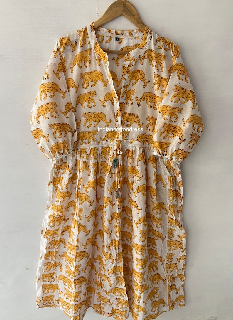 May include: White cotton dress with a yellow tiger print. The dress has a V-neckline, 3/4 sleeves, and a drawstring waist. The dress is made in India and is available in sizes S-XL.