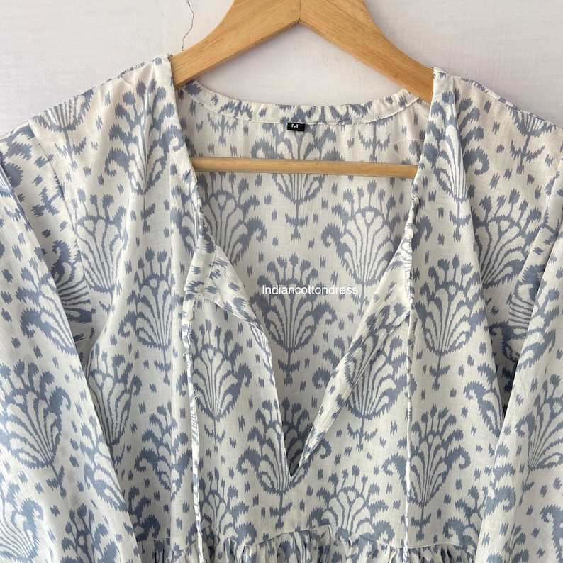 May include: A white cotton dress with a blue ikat print and a V-neckline. The dress has a tie at the neckline and long sleeves.