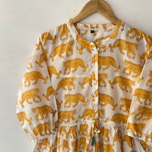 May include: A white cotton dress with a mandarin collar and long sleeves. The dress is covered in a repeating pattern of yellow tigers. The dress has a drawstring waist and a tassel.