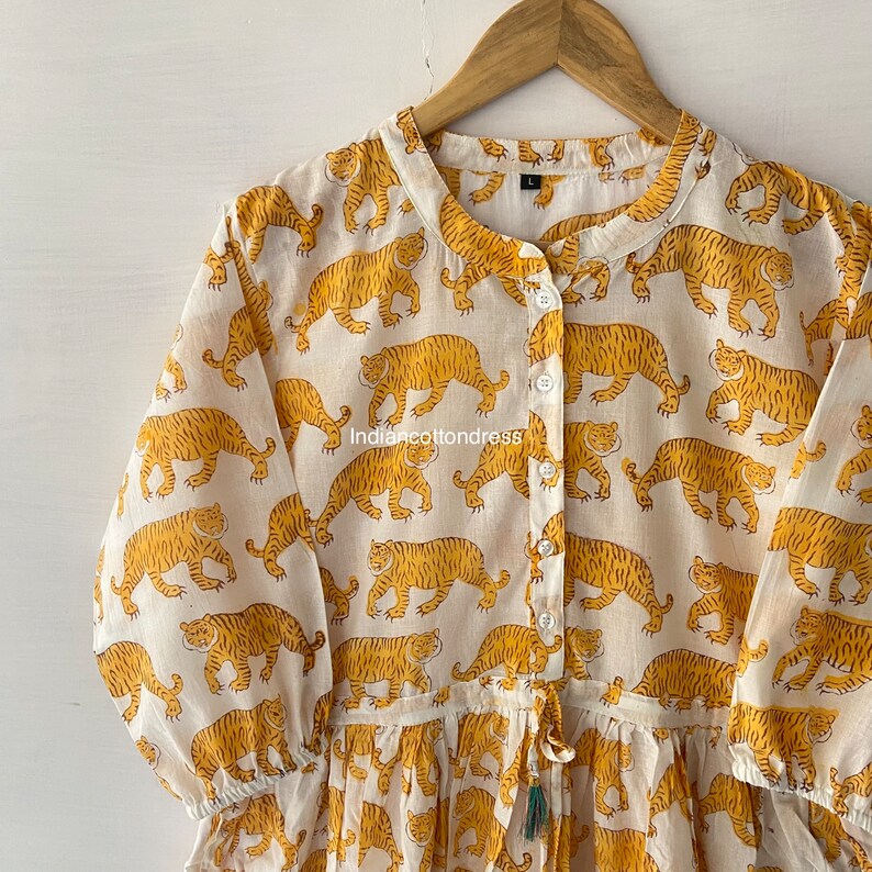 May include: A white cotton dress with a mandarin collar and long sleeves. The dress is covered in a repeating pattern of yellow tigers. The dress has a drawstring waist and a tassel.