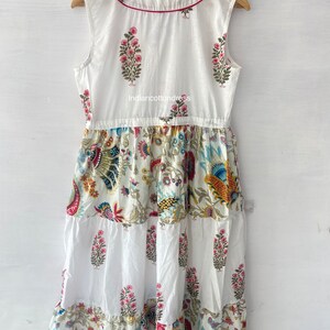 May include: White cotton dress with a floral print. The dress has a tiered skirt and a sleeveless bodice. The dress is decorated with a red trim around the neckline.