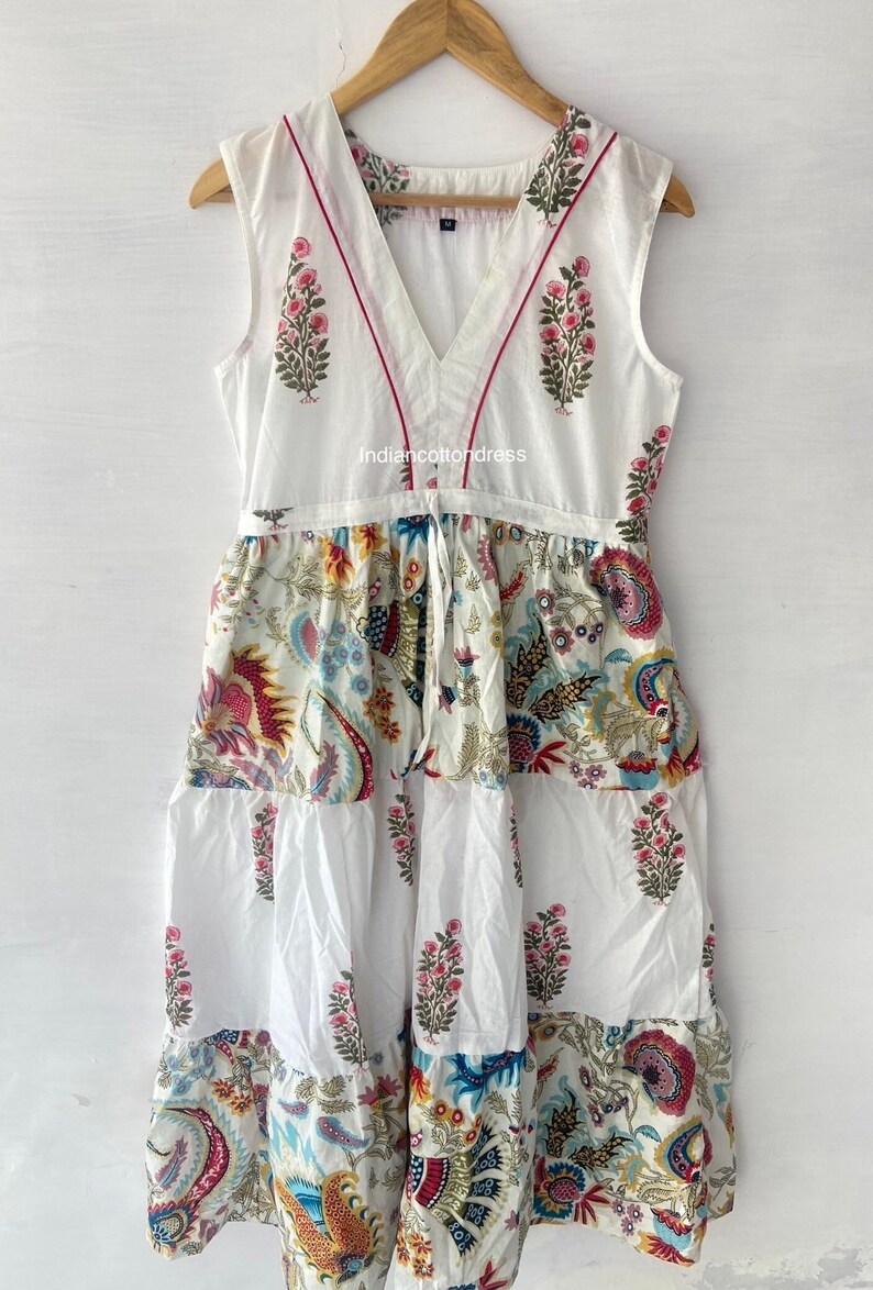 May include: White cotton dress with a V-neckline and a tie at the waist. The dress has a floral print in shades of pink, blue, yellow, and green. The dress is sleeveless and has a tiered skirt.