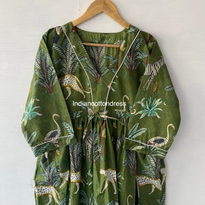 May include: A green cotton dress with a V-neckline and white trim. The dress features a jungle print with leopards, zebras, flamingos, and palm trees. The dress has a gathered waist and 3/4 sleeves.
