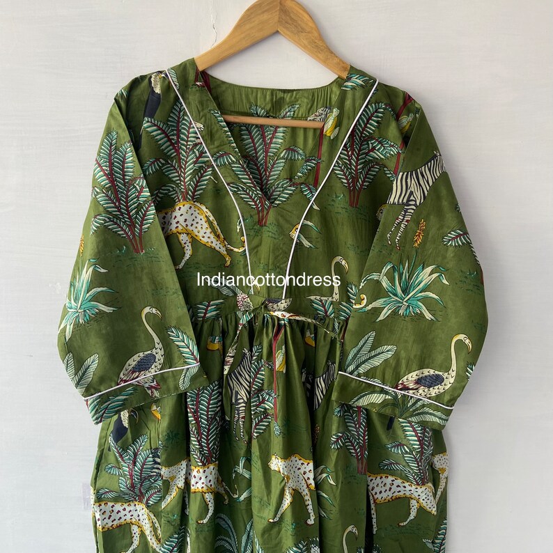May include: A green cotton dress with a V-neckline and white trim. The dress features a jungle print with leopards, zebras, flamingos, and palm trees. The dress has a gathered waist and 3/4 sleeves.
