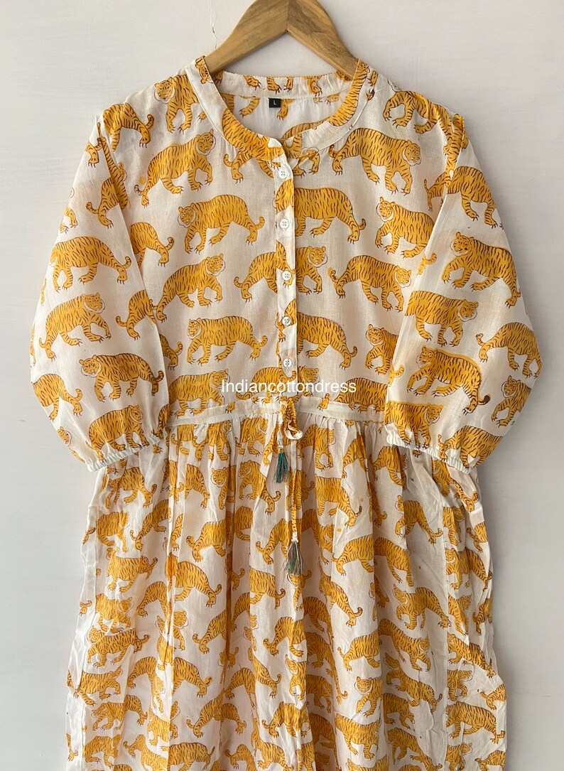 May include: A white cotton dress with a yellow tiger print. The dress has a button-up front, long sleeves, and a drawstring waist.