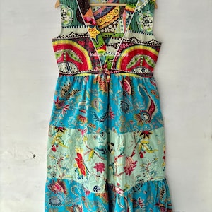 May include: A long, sleeveless, patchwork dress with a V-neckline. The dress is made of colorful cotton fabric with floral and geometric patterns. The dress has a tiered skirt and a tie at the waist. The text "Indian Cotton Dress" is visible on the dress.