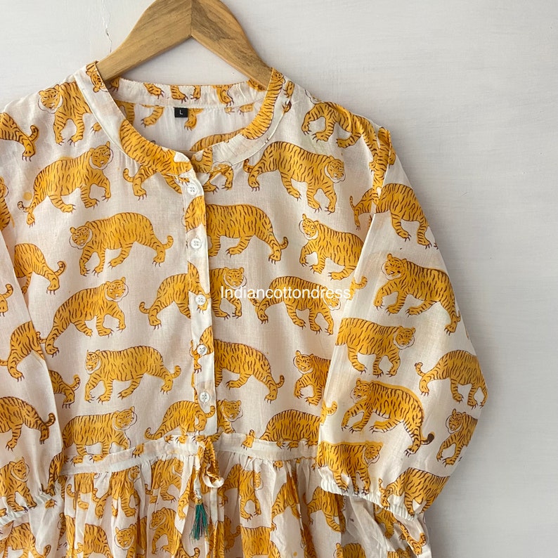 May include: A white cotton dress with a yellow tiger print. The dress has a button-up front, long sleeves, and a drawstring waist.