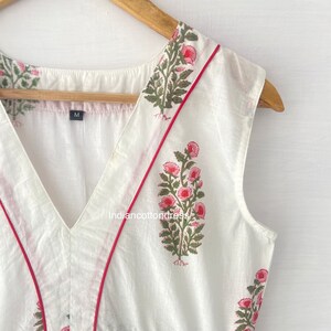 May include: White sleeveless top with a V-neckline and a pink floral print. The neckline and armholes are trimmed with a red piping. The top has a size tag that says "M".
