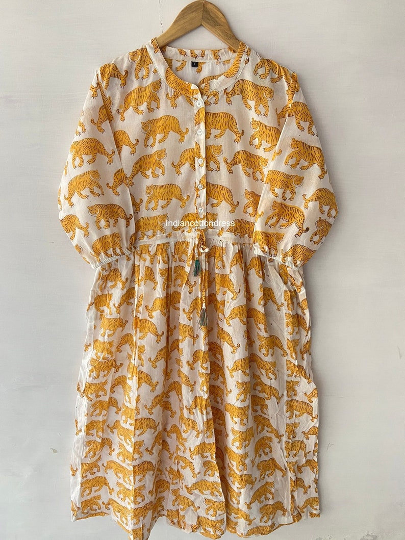 May include: A white cotton dress with a yellow tiger print. The dress has a button-up front and a drawstring waist. The dress is 3/4 sleeve and has a relaxed fit.