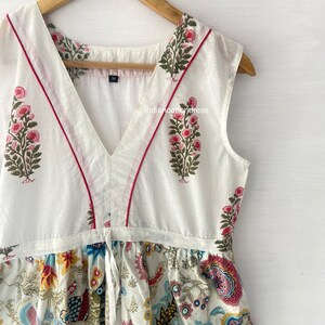 May include: White sleeveless dress with a V-neckline and a floral print. The dress has a tie at the waist and a red trim around the neckline.