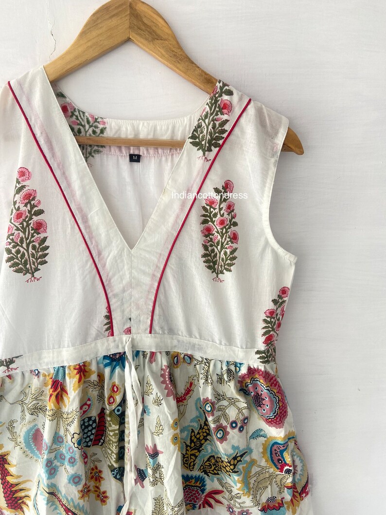 May include: White sleeveless dress with a V-neckline and a floral print. The dress has a tie at the waist and a red trim around the neckline.