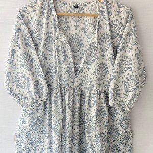 May include: A white cotton dress with a blue floral print and a V-neck. The dress has a tie at the neckline and 3/4 sleeves.