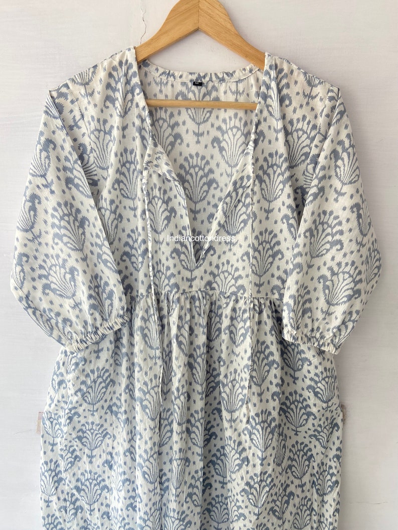 May include: A white cotton dress with a blue floral print and a V-neck. The dress has a tie at the neckline and 3/4 sleeves.