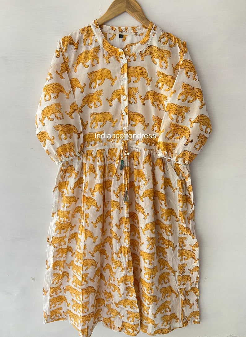 May include: A white cotton dress with a yellow tiger print. The dress has a button-up front, 3/4 sleeves, and a drawstring waist.
