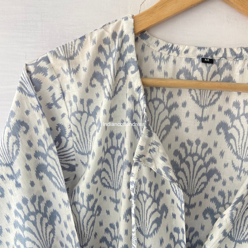May include: A white cotton shirt with a blue ikat pattern. The shirt has a V-neck and a button-up front. The shirt is hanging on a wooden hanger. The size tag says "M".