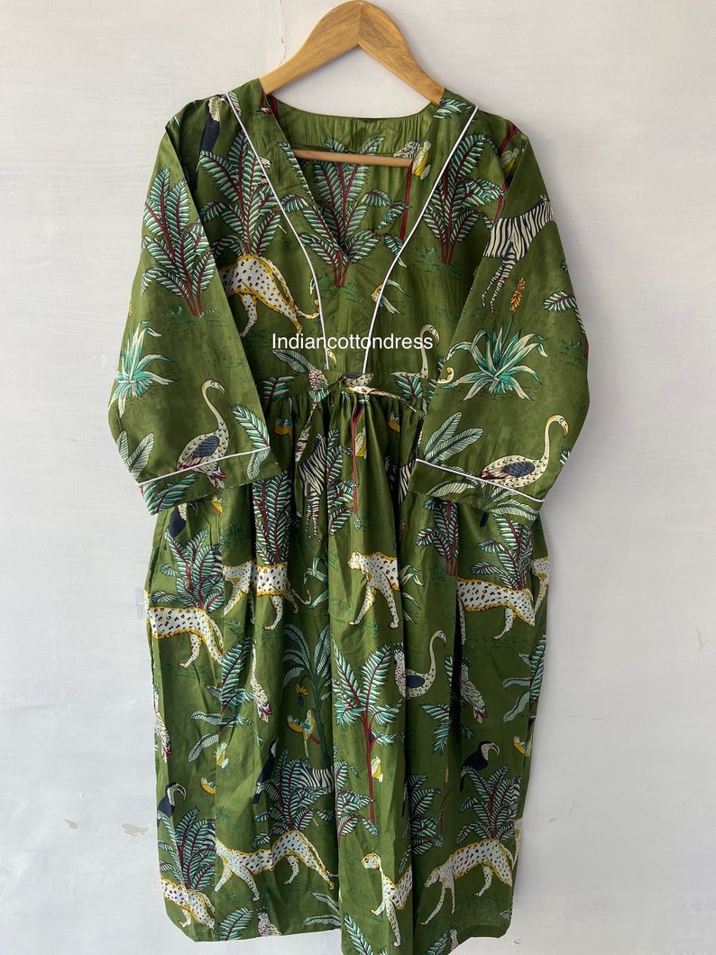 May include: A green cotton dress with a white trim and a jungle print featuring leopards, zebras, birds, and palm trees. The dress has a V-neckline and a tie at the waist. The text "Indiancottondress" is visible on the dress.
