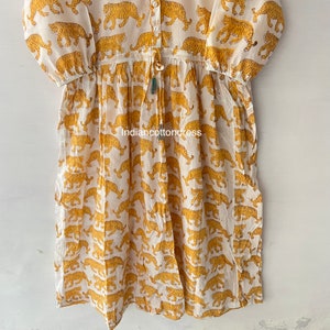 May include: A white cotton dress with a yellow tiger print. The dress has a button-up front, a drawstring waist, and 3/4 sleeves.