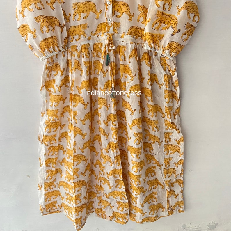 May include: A white cotton dress with a yellow tiger print. The dress has a button-up front, a drawstring waist, and 3/4 sleeves.