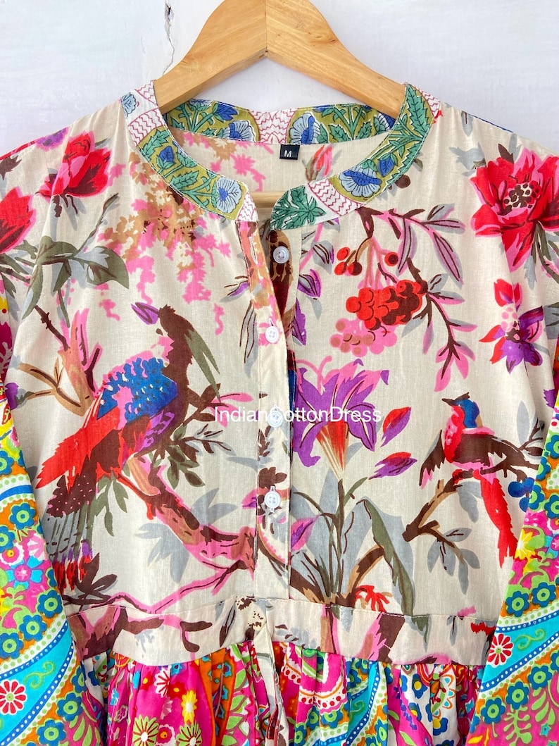 May include: A beige cotton dress with a floral print featuring birds and flowers. The dress has a button-up front and a ruffled hem. The sleeves are long and have a colorful paisley print. The dress has a size tag that says "M".