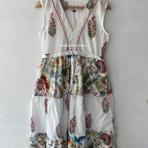 May include: White sleeveless dress with a V-neckline and a tiered skirt. The dress has a floral print in shades of pink, blue, and yellow. The neckline is trimmed with a red ribbon. The dress is made of cotton and is perfect for summer.
