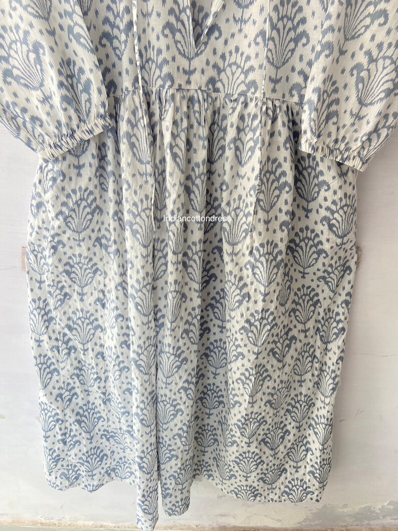May include: A white cotton dress with a blue floral pattern. The dress has a V-neckline and 3/4 sleeves. The dress is gathered at the waist and has a flowy skirt.