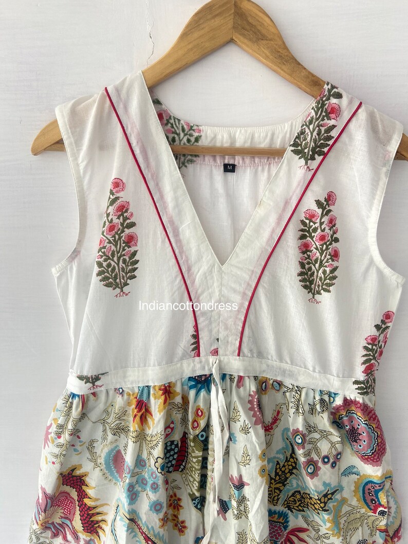 May include: White sleeveless dress with a V-neckline and a floral print. The dress has a tie at the waist and a red trim around the neckline. The dress is made of cotton and is available in size M.