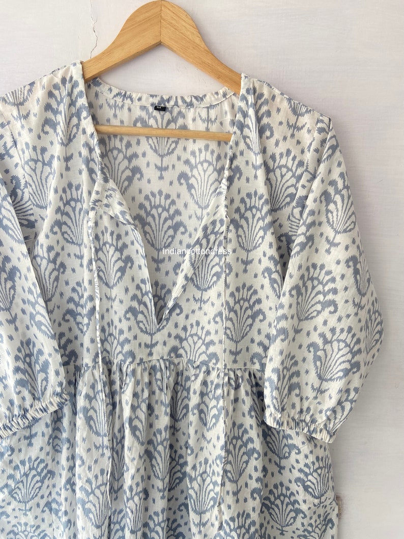 May include: A white cotton dress with a blue floral print and a V-neckline. The dress has a tie at the neckline and 3/4 sleeves.