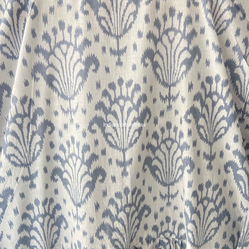 May include: Close-up of a white fabric with a blue floral pattern. The fabric has a subtle texture and a slightly distressed look. The pattern is a repeating design of stylized flowers with a central stem and multiple petals.