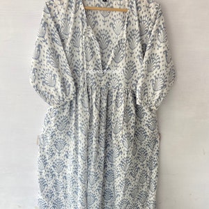May include: A white cotton dress with a blue floral print and a V-neckline. The dress has a gathered skirt and 3/4 sleeves. The dress is hanging on a wooden hanger.