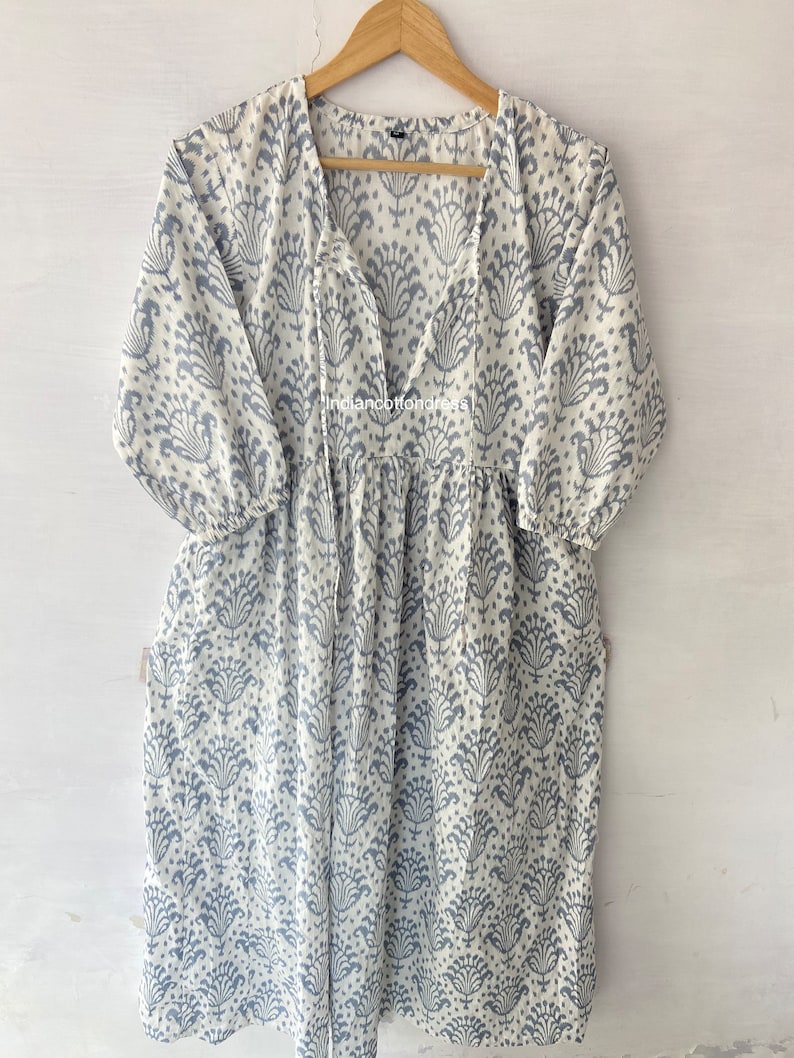 May include: A white cotton dress with a blue floral print and a V-neckline. The dress has a gathered skirt and 3/4 sleeves. The dress is hanging on a wooden hanger.