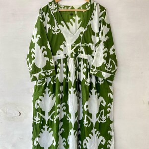 May include: A green and white cotton dress with a V-neck and 3/4 sleeves. The dress has a white ikat pattern on a green background. The dress is hanging on a wooden hanger.