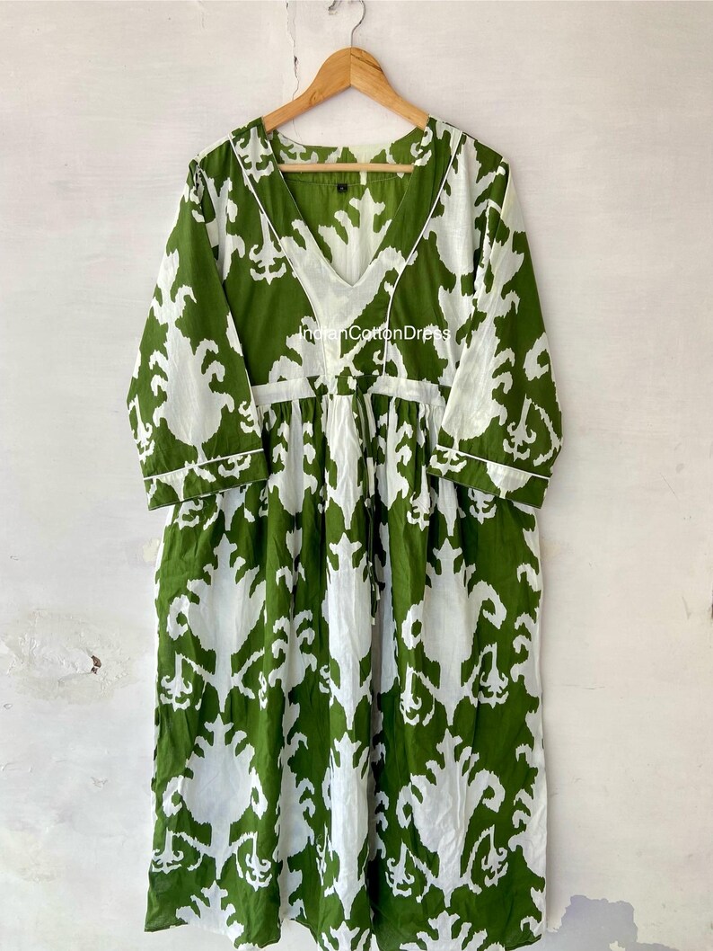 May include: A green and white cotton dress with a V-neck and 3/4 sleeves. The dress has a white ikat pattern on a green background. The dress is hanging on a wooden hanger.