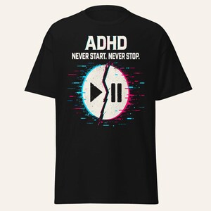 Op de afbeelding: Een zwart t-shirt met de witte tekst ADHD NEVER START. NEVER STOP. boven een cirkelvormige afbeelding. De cirkel is gespleten door een grillige lijn, met een afspeelpictogram links en een pauzepictogram rechts, voorzien van blauwe en roze glitch-effecten.