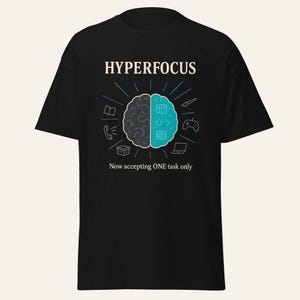 Op de afbeelding: Een zwart t-shirt met een afbeelding van een gespleten brein, met de linkerkant in grijs en de rechterkant in blauwgroen. De tekst HYPERFOCUS staat erboven en Now accepting ONE task only eronder, omringd door kleine pictogrammen.