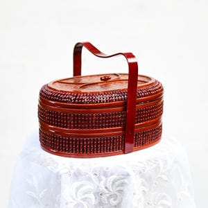 May include: A red, oval-shaped, tiered basket with a handle. The basket has a textured surface with a repeating pattern of small, raised dots. The handle is arched and attached to the sides of the basket. The basket is sitting on a white, lacy tablecloth.
