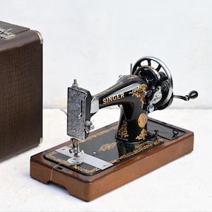 Op de afbeelding: Een antieke Singer naaimachine met een zwarte en gouden afwerking, geplaatst op een houten basis. De machine heeft ingewikkelde gouden details en een handslinger. Een bruine draagtas is links zichtbaar.