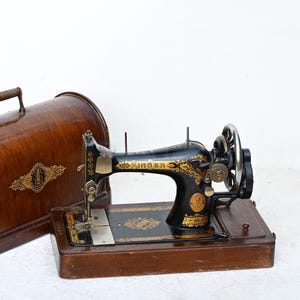 May include: An antique Singer sewing machine with a black and gold finish, displayed next to a wooden carrying case. The machine features intricate gold detailing and a large hand wheel. The wooden case has a handle and decorative gold accents.