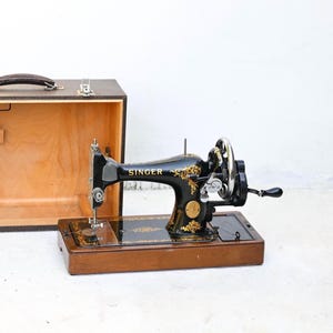 May include: A vintage Singer sewing machine in black with gold floral accents, set on a wooden base. The machine features a hand crank and is accompanied by a wooden carrying case with a handle.